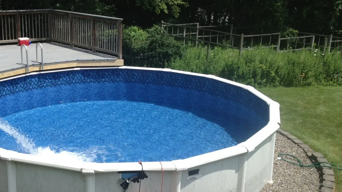 A round above-ground pool beside a wood deck with a ladder, lawn and trees behind