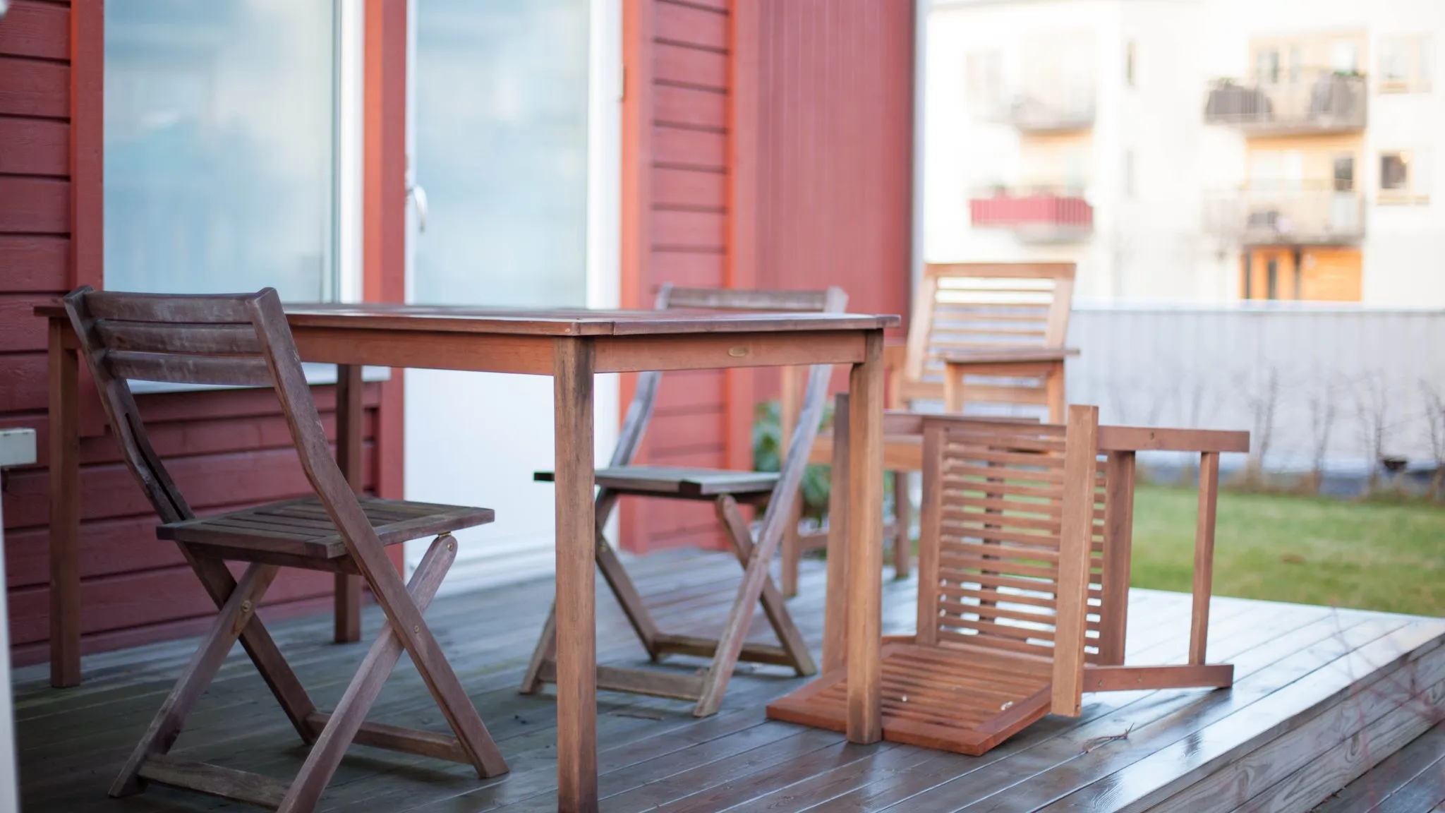 A wooden backyard deck with a folding table and chairs against a red house, one chair folded flat — the furniture a big deck box exists to put away