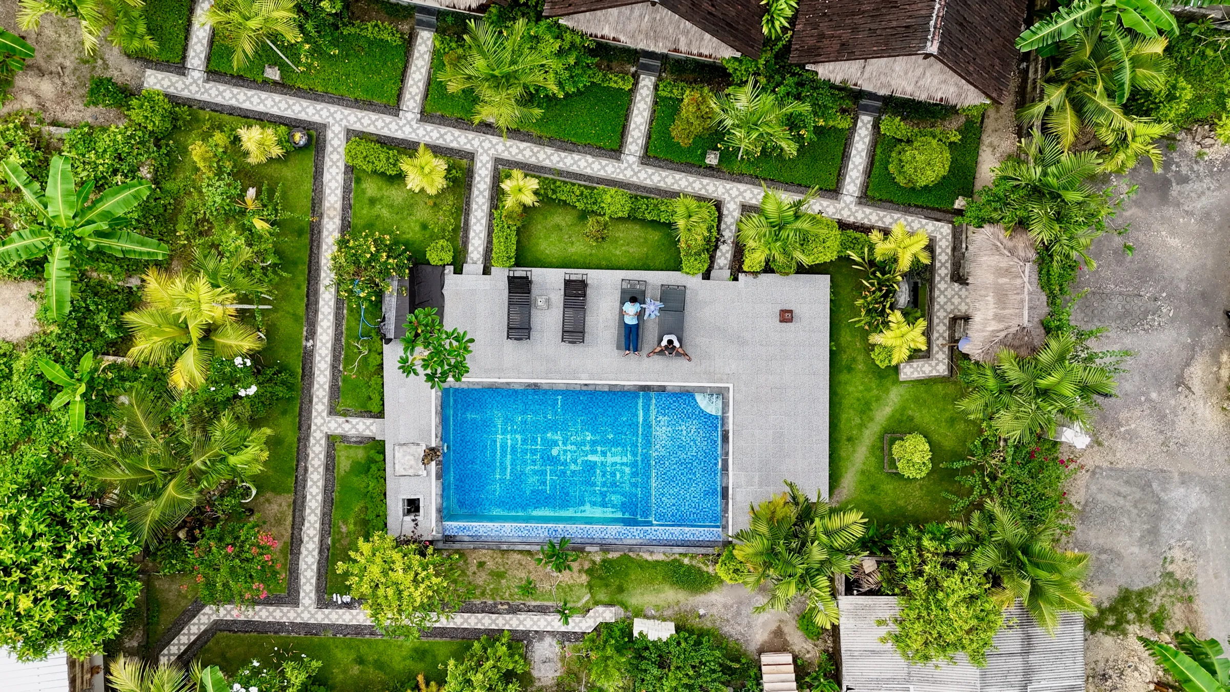 Aerial view of a bright blue pool at a tropical cottage, boxed in by lush greenery with a lounger poolside