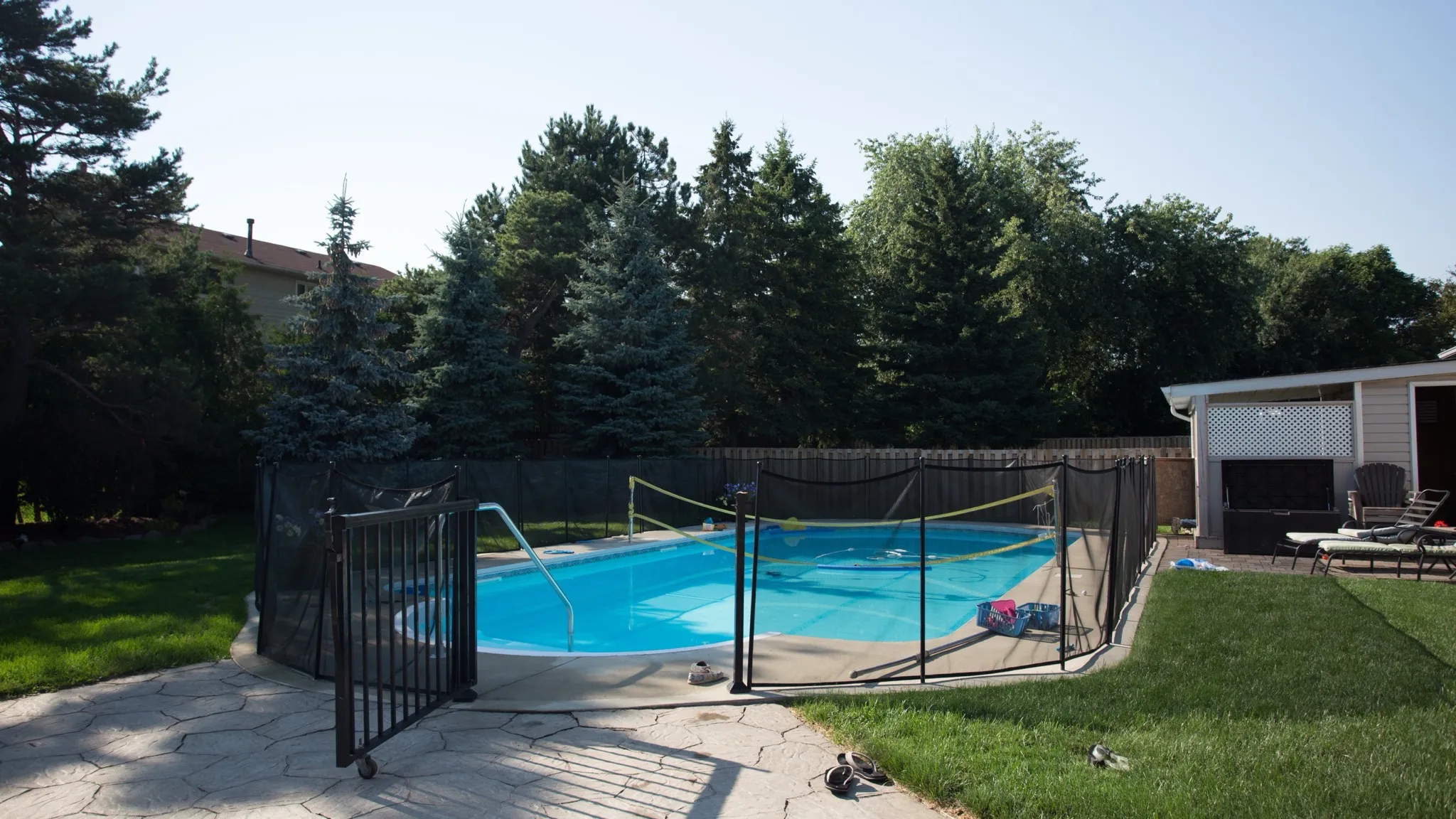 A backyard in-ground pool behind a black mesh safety fence, with a stamped-concrete deck in front and evergreens behind
