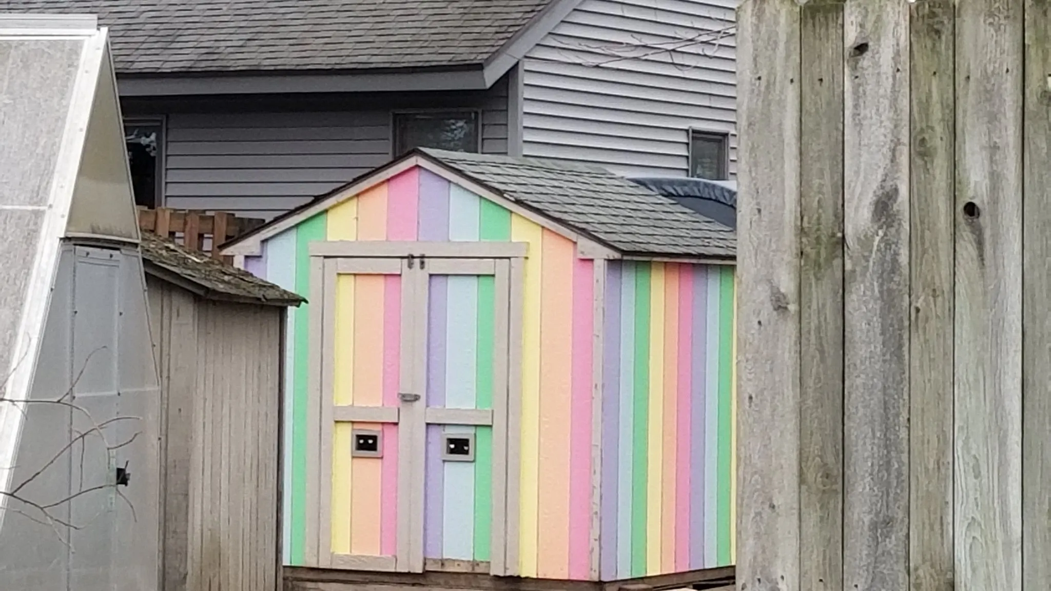 A small backyard storage shed painted in pastel rainbow stripes beside a weathered wooden fence, a grey house behind it — the corner of a yard a shed goes in