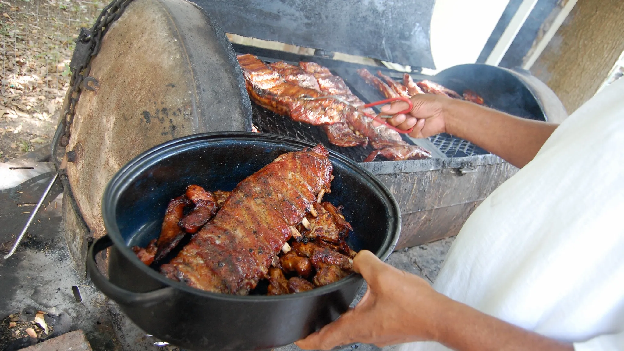 Racks of spare ribs on the open grate of a welded barrel smoker pit, with a cook holding a pan of finished ribs in the foreground