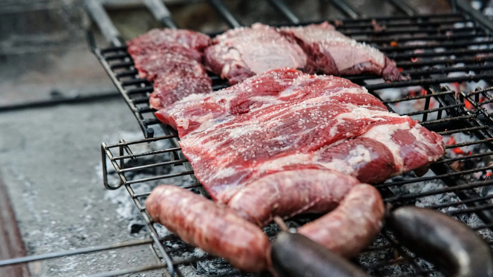 Raw salted steaks and sausages laid across a grill grate over glowing coals, the grate filling the frame