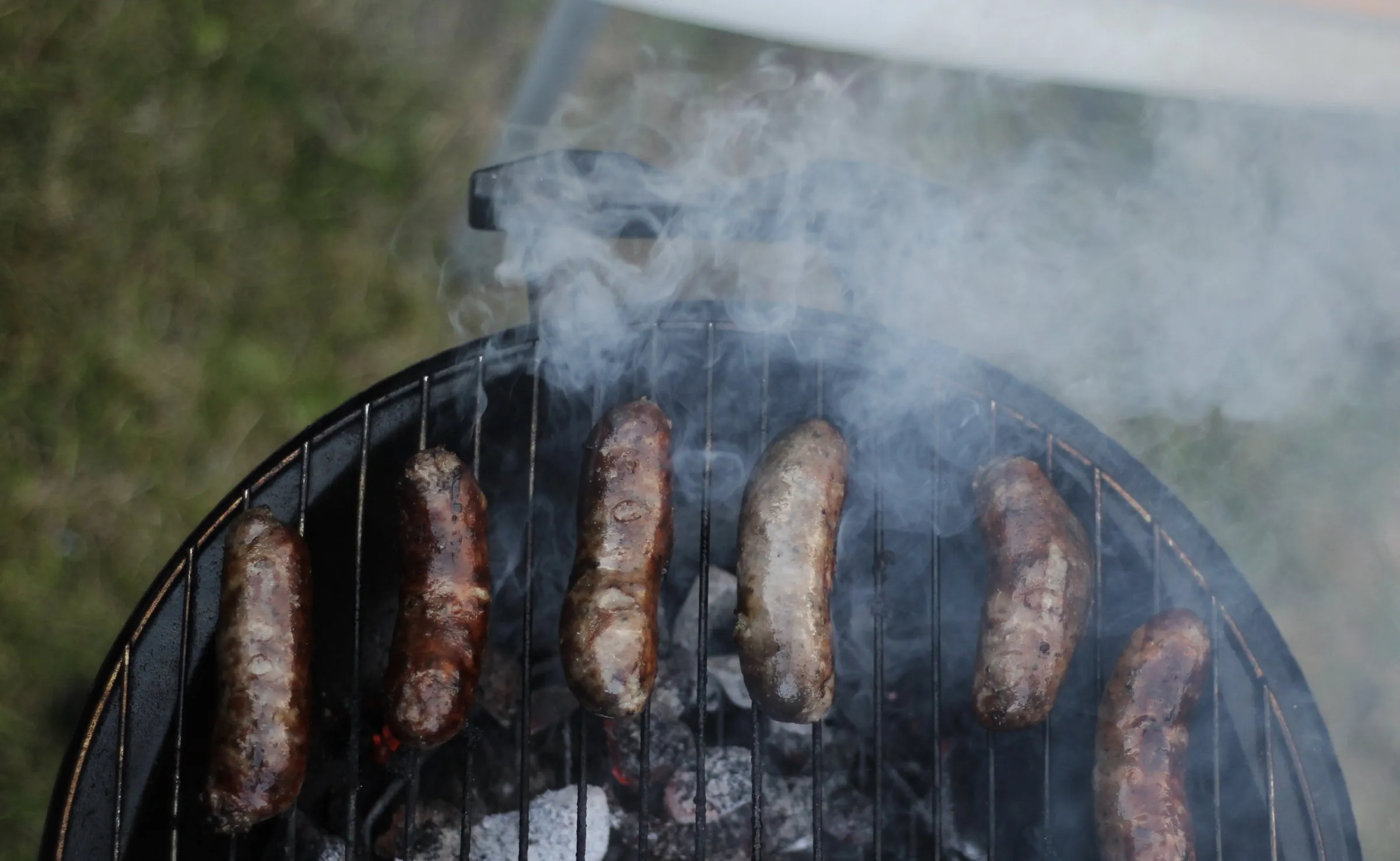Sausages on the grate of a round charcoal cooker with smoke rising off the coals