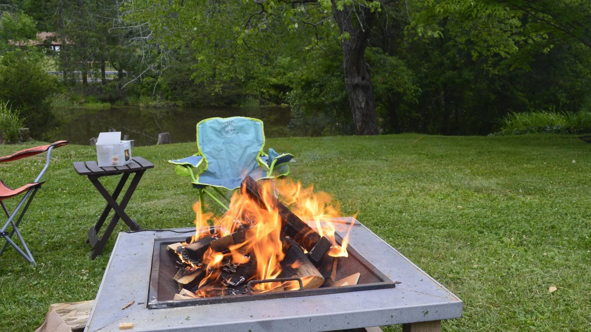 A square steel wood fire pit burning hard on a lawn beside a pond, two folding camp chairs and a small side table pulled up to it