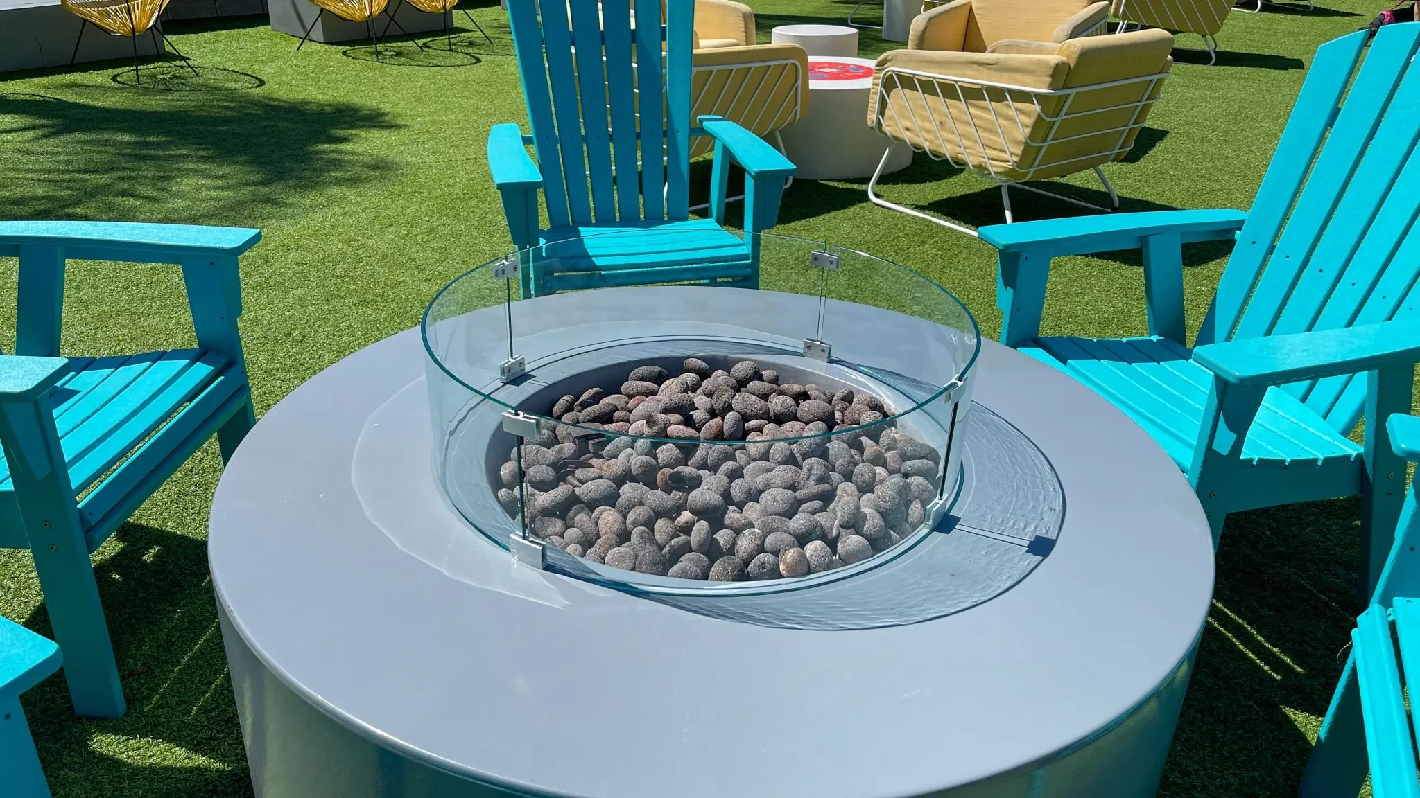 A round grey propane fire table with lava rock and a glass wind guard ringed by turquoise Adirondack chairs on a lawn, yellow patio chairs behind