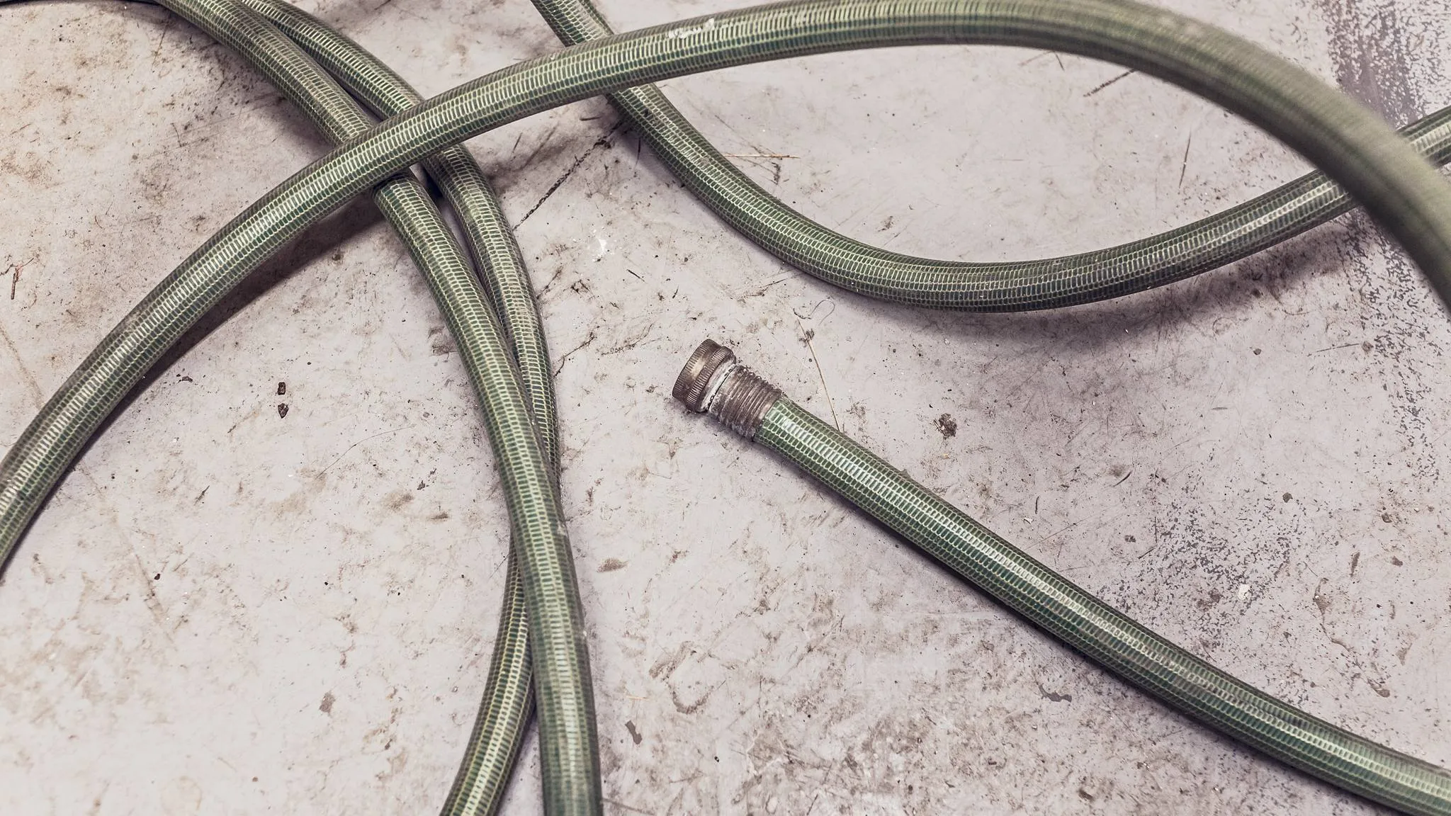 A loose green garden hose looping across a scuffed concrete floor, its brass coupling in the foreground