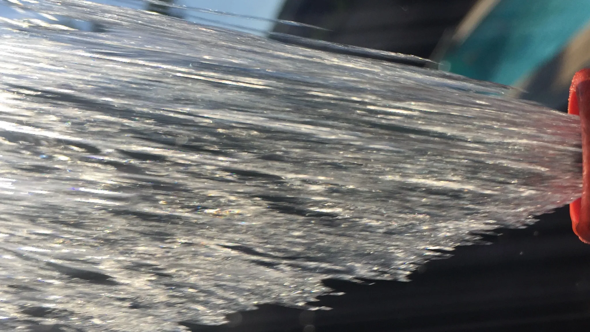A fan of water spraying from the end of a garden hose nozzle over a lawn, backlit by low sun