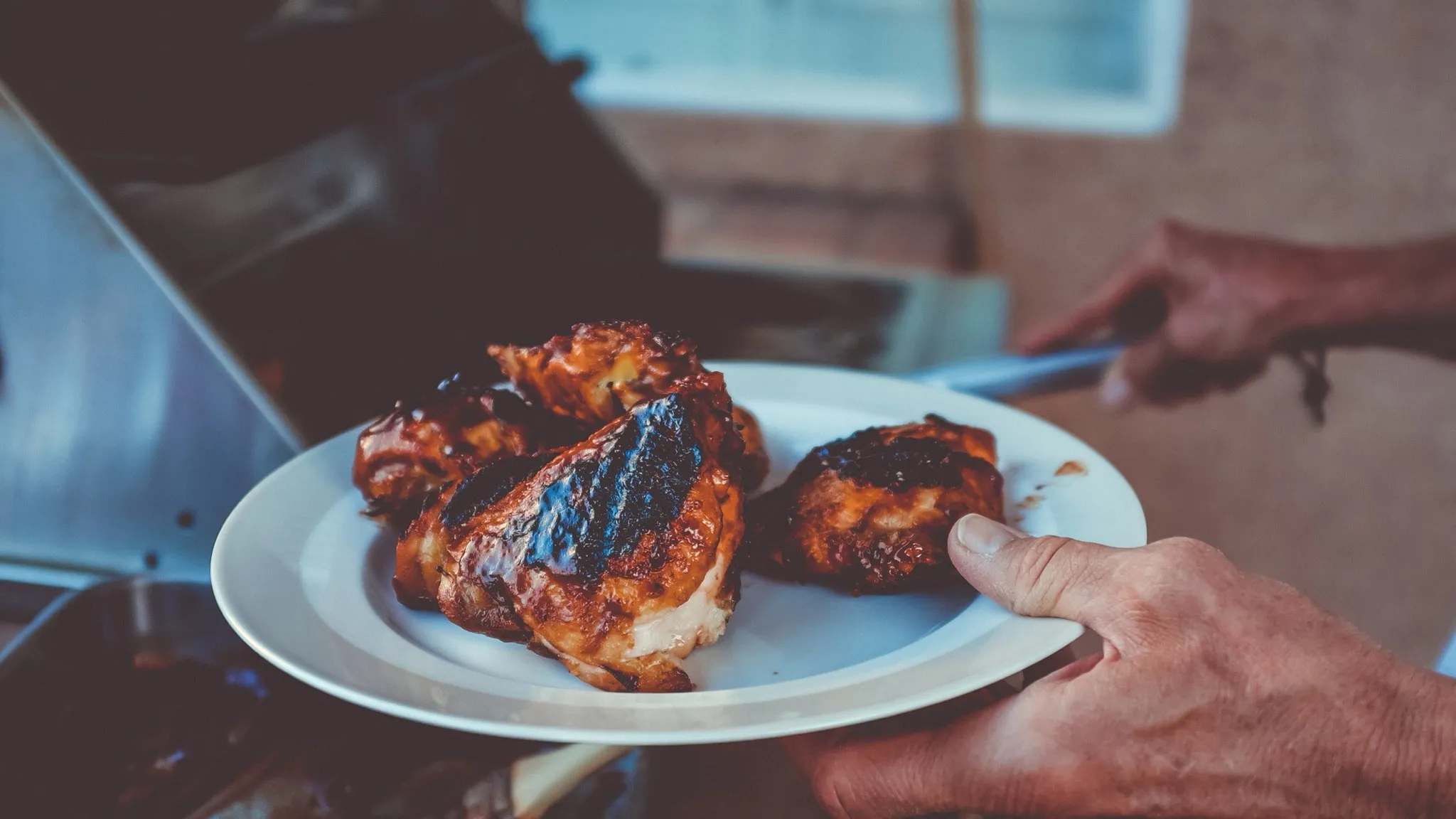 A hand holding a plate of barbecue-glazed chicken just lifted off a stainless gas grill, the grill's lid and grate out of focus behind it