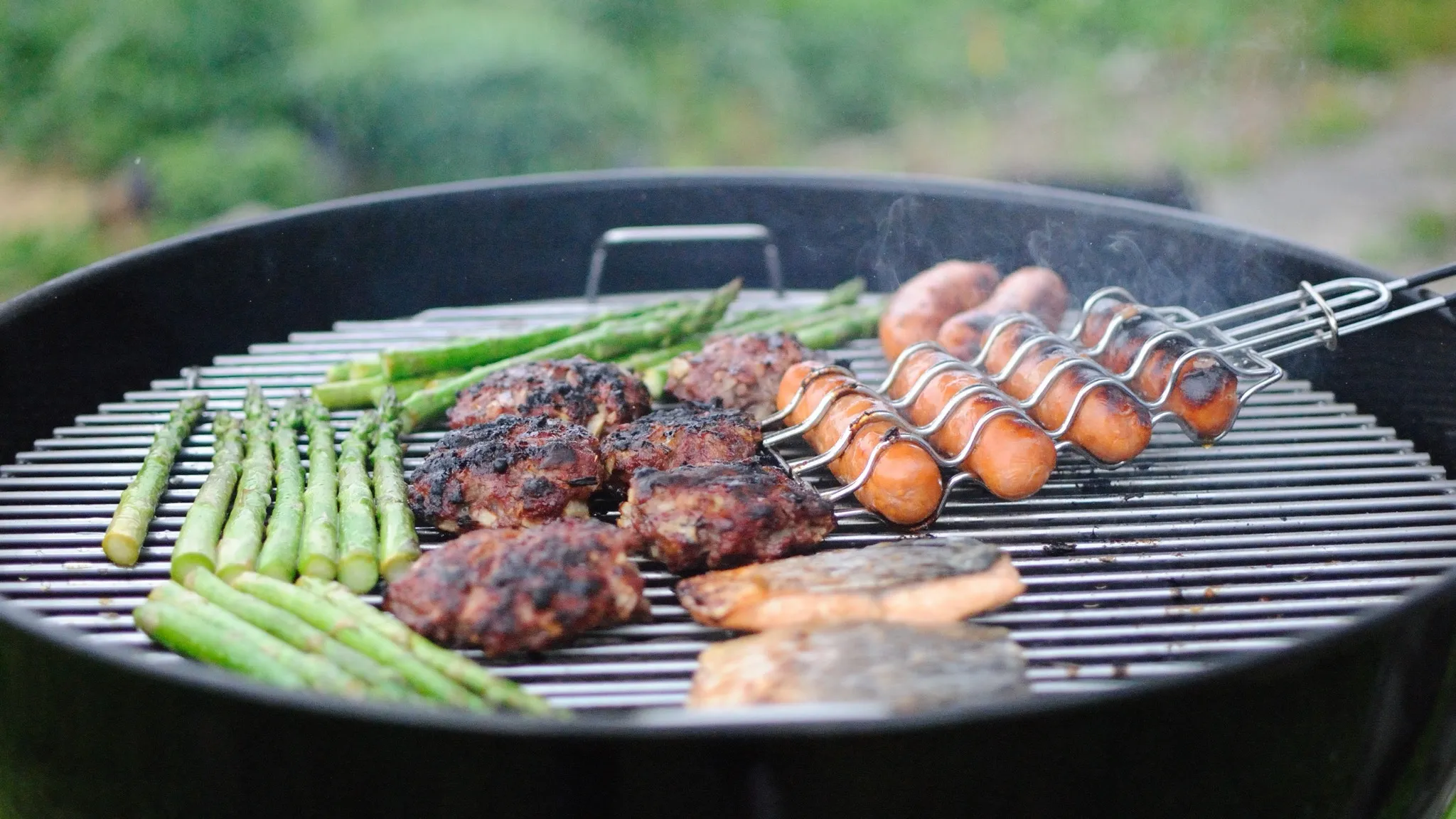 Burgers, asparagus and sausages on a round kettle grill outdoors, a garden blurred behind