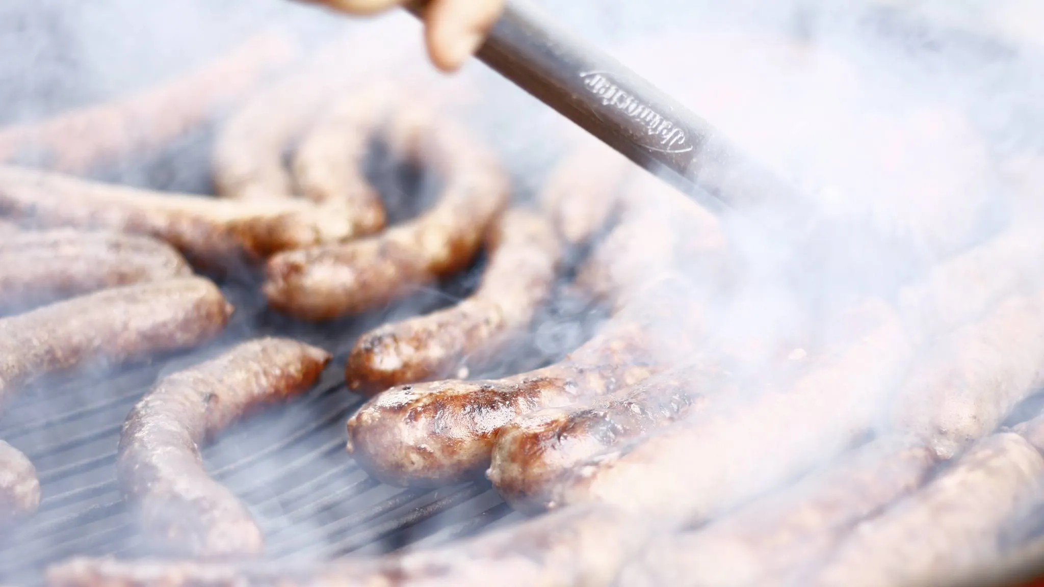 Sausages browning on a grill grate in heavy smoke while tongs turn them