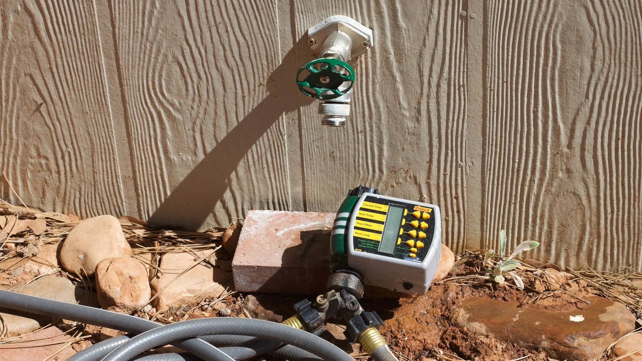 An older digital hose timer, unhooked for the season, lies on a brick beneath an outdoor spigot with a green handle, a grey garden hose coiled in front of it