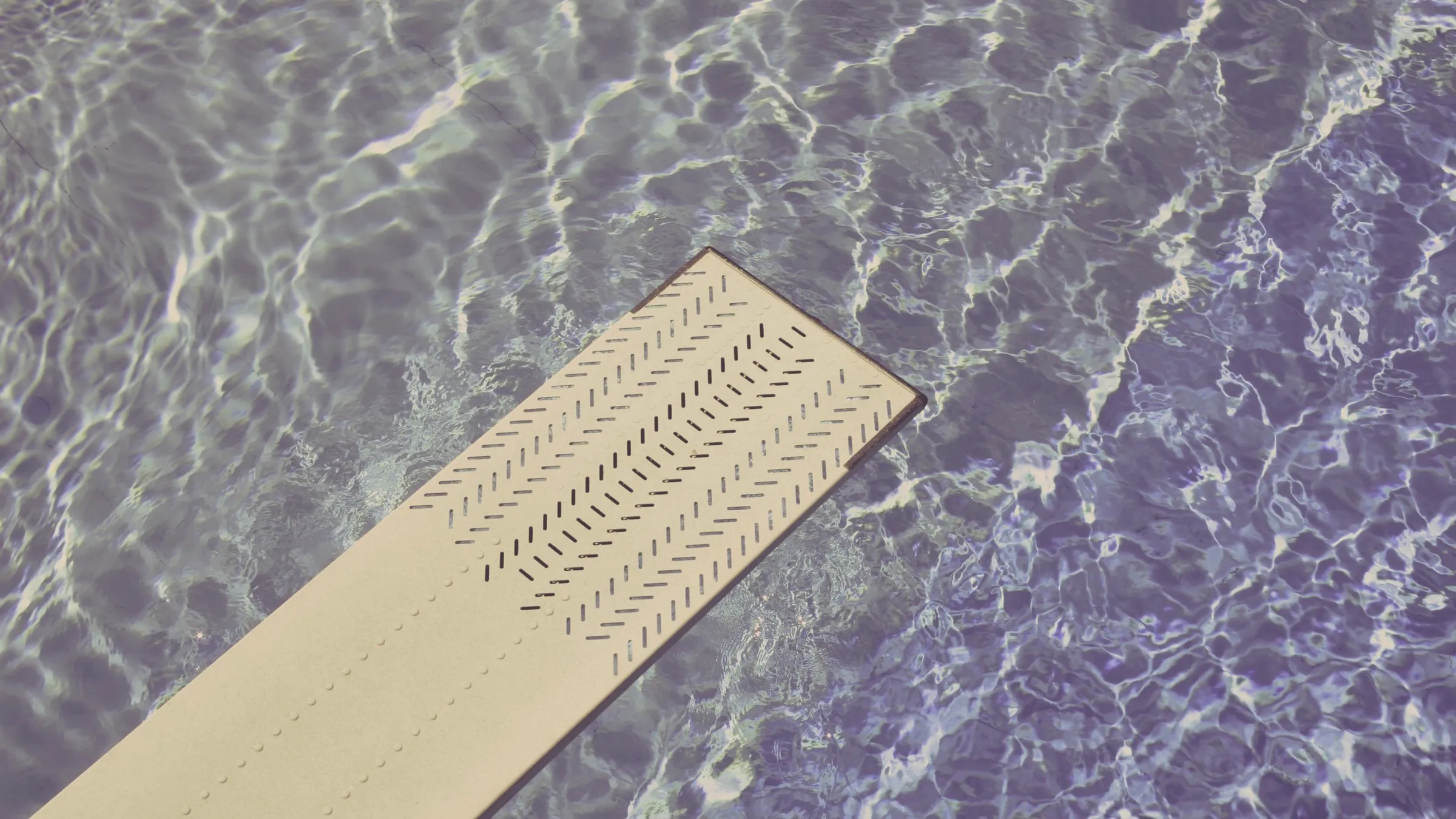 Looking down a diving board over the clear, sunlit rippling water of an in-ground swimming pool