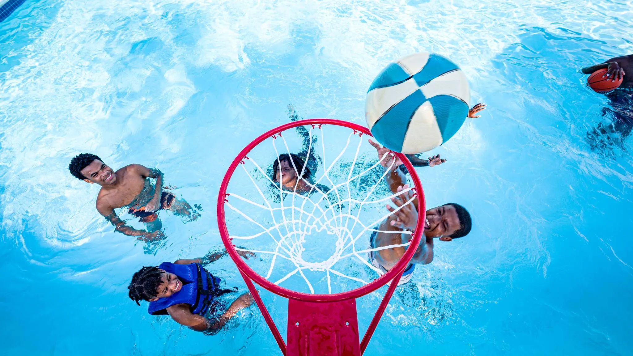 Looking straight down over a red pool basketball rim as a ball drops toward it and kids in the water reach up