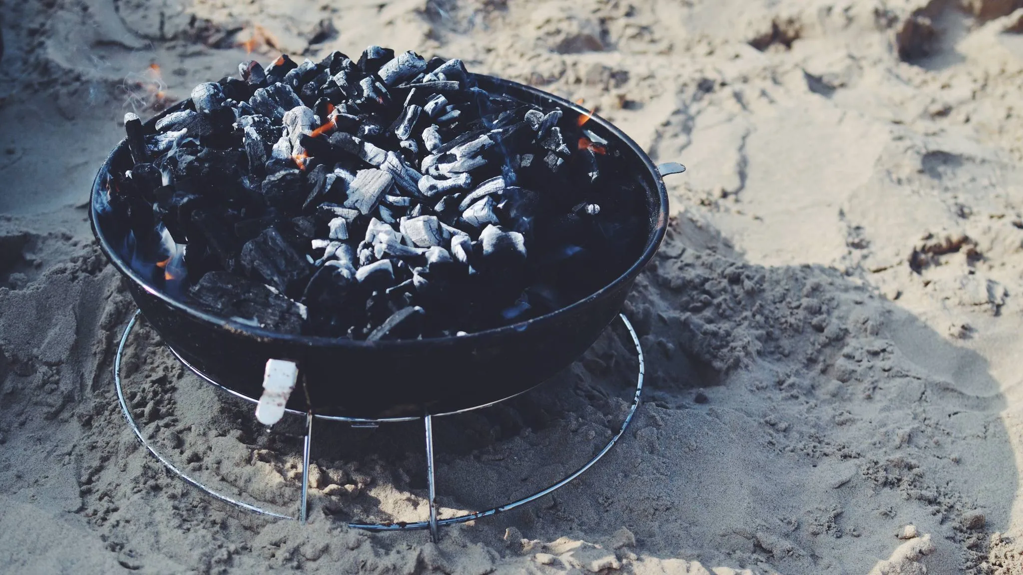 A small black charcoal bowl grill on a wire ring, full of lit lump charcoal, sitting in beach sand