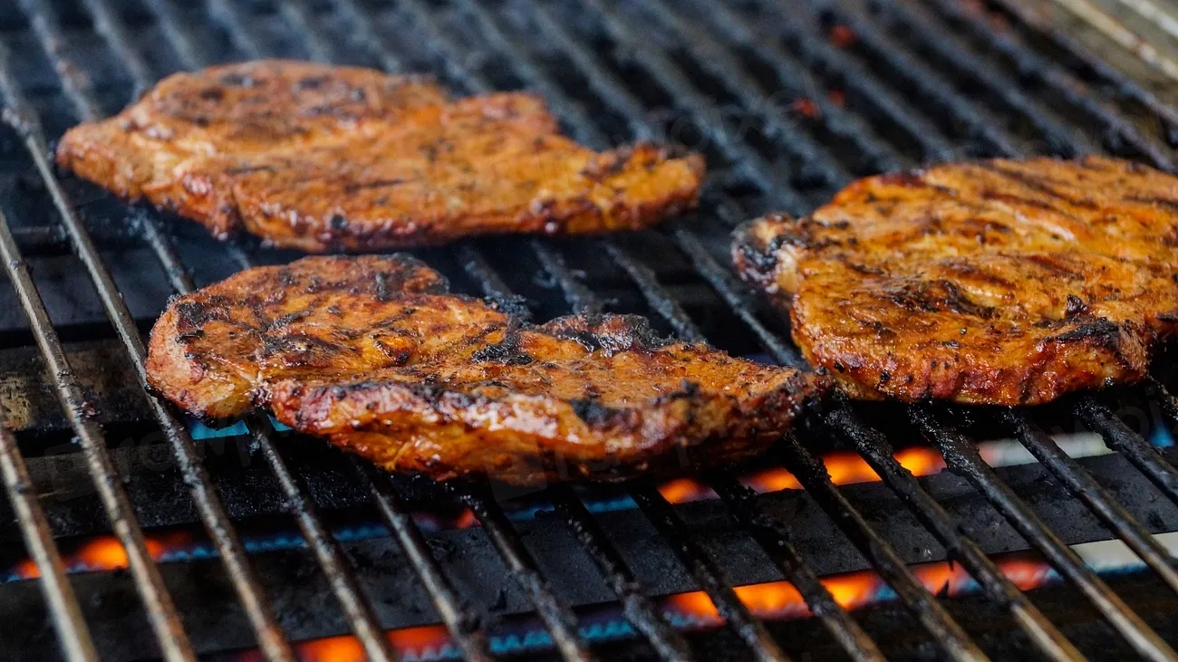 Seasoned pork steaks searing on a stainless gas grill grate, with blue and orange burner flame visible beneath the bars