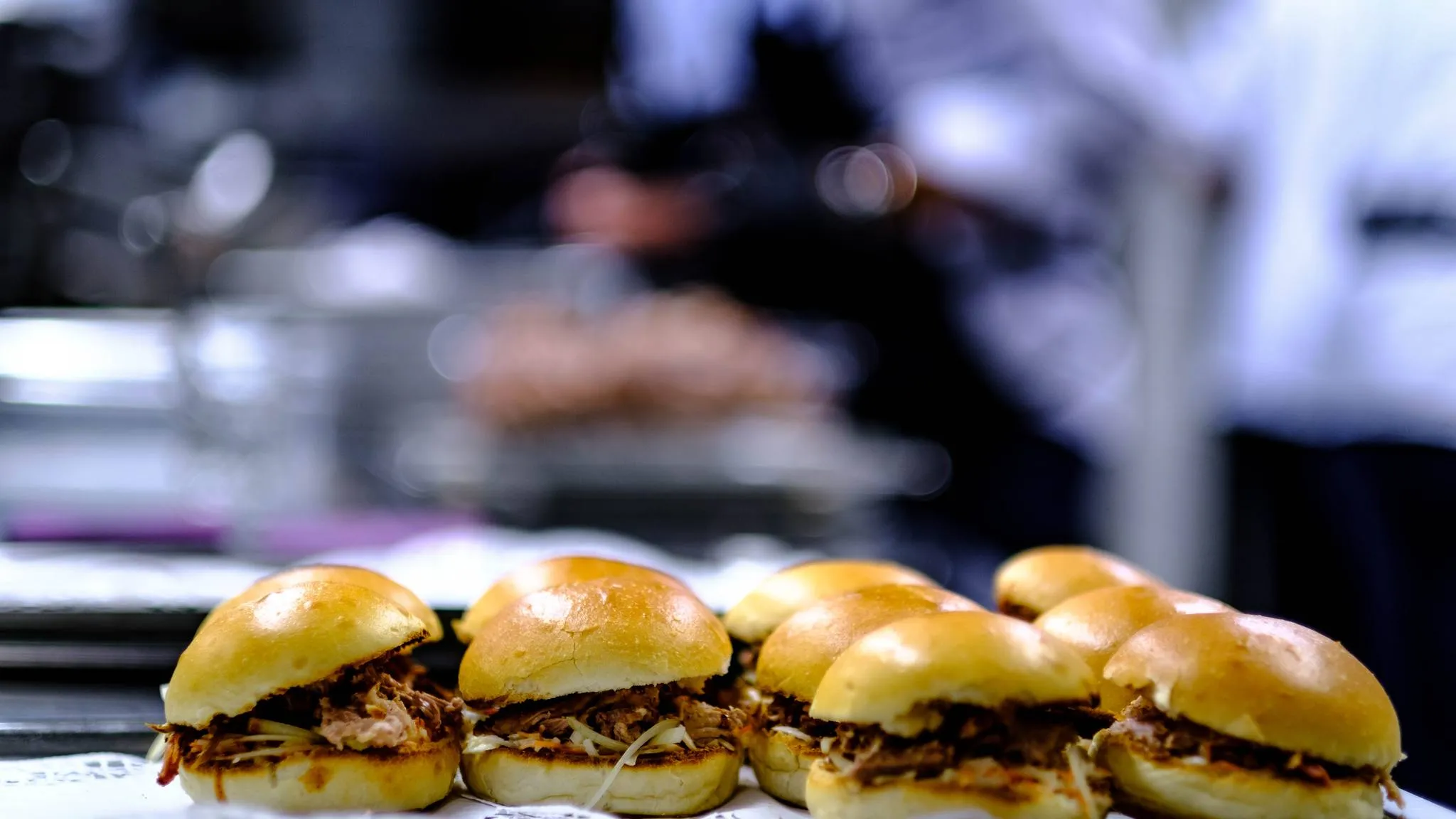 A tray of pulled pork sliders on soft buns with a cook blurred in the background