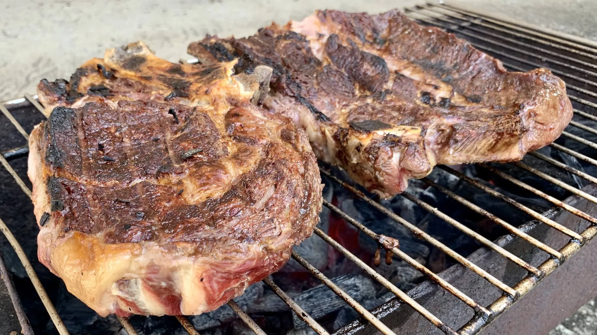 Two seared rib steaks on a grill grate over glowing embers