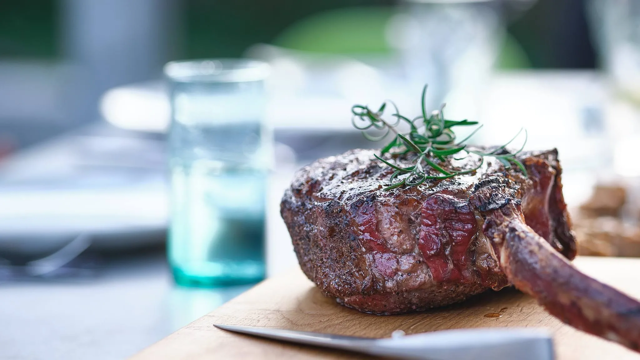 A smoked tomahawk steak topped with rosemary on a wooden board at an outdoor table, a water glass out of focus behind it