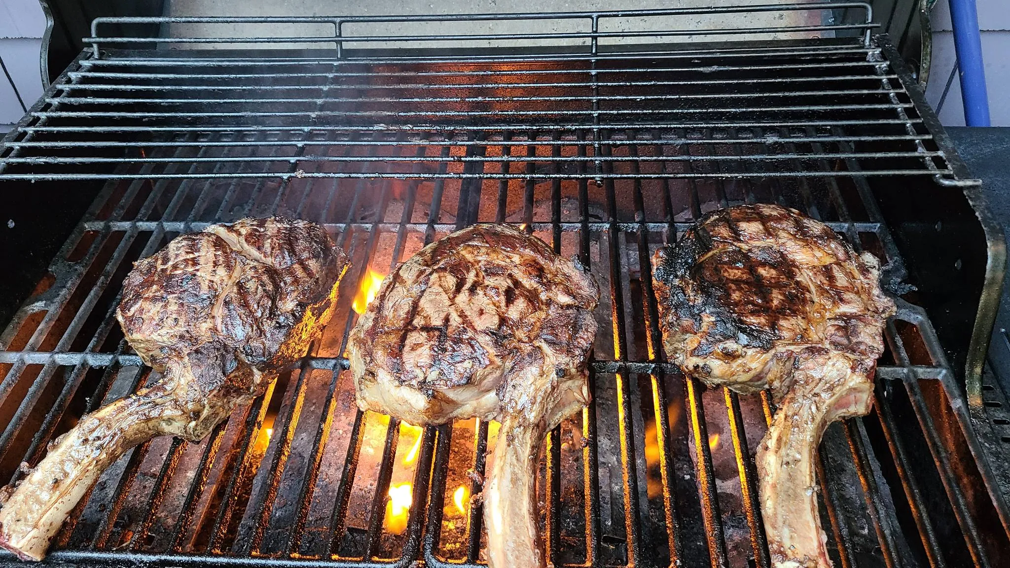 Three tomahawk steaks searing over open flame on a gas grill grate, a warming rack above