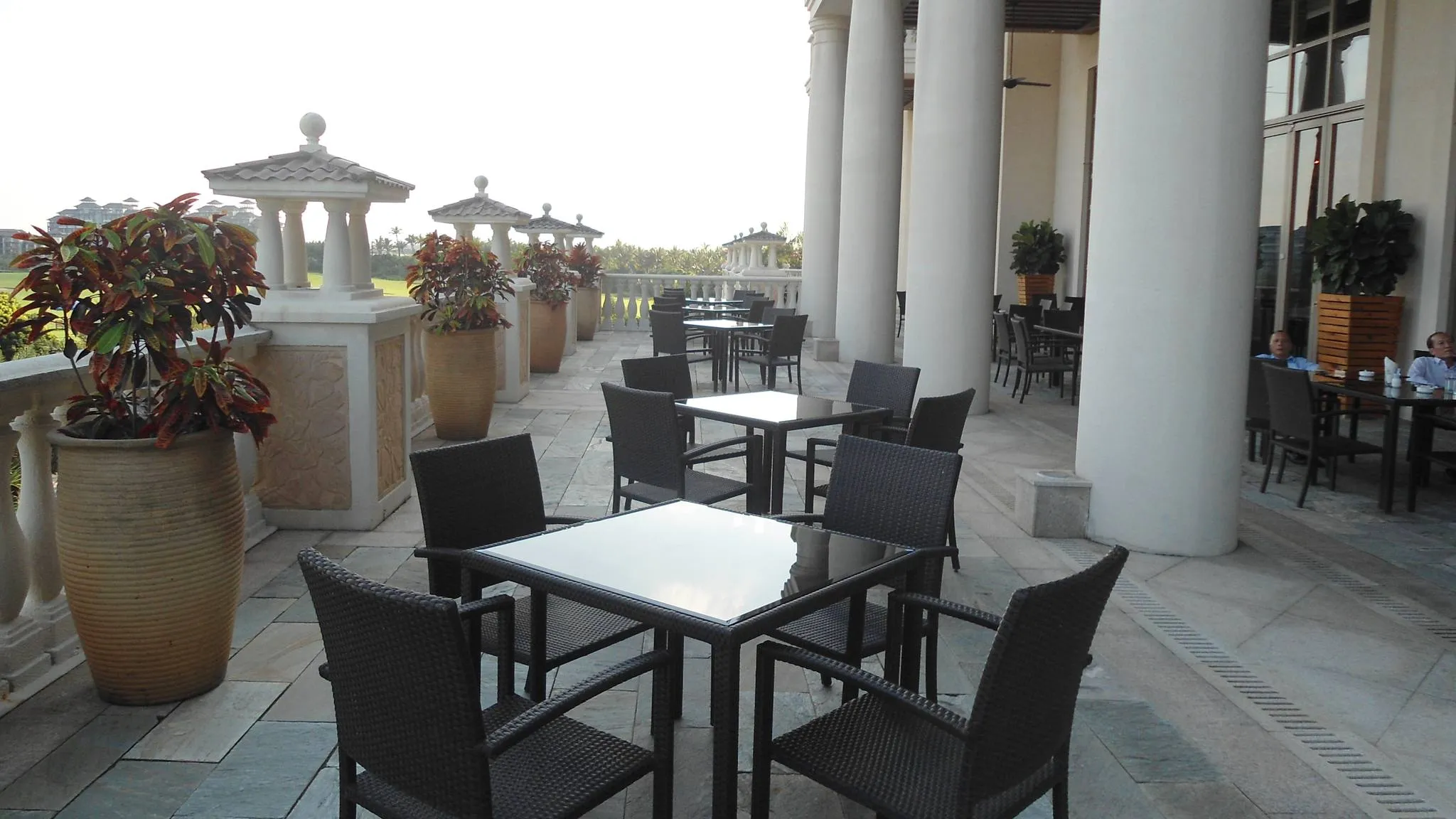 Dark wicker armchairs around glass-topped tables on a stone hotel terrace with white columns