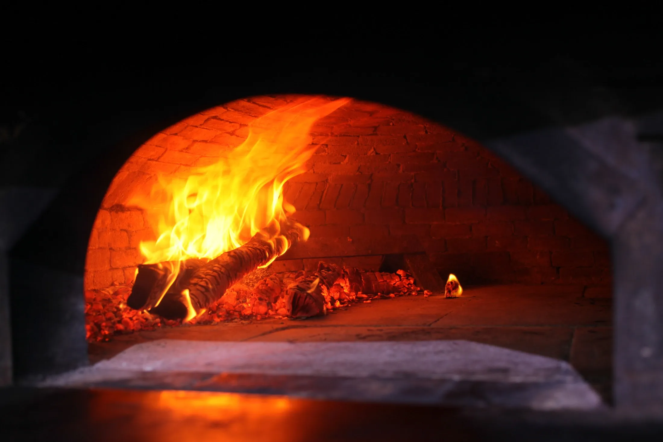 Live flames burning inside a brick wood-fired oven against the dark — live-fire cooking ambiance
