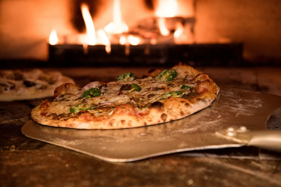 A fresh wood-fired pizza on a metal peel in front of the flames of a pizza oven